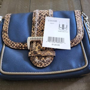 May include: A blue handbag with a snakeskin pattern trim and strap. The bag has a silver-colored buckle and a tag that reads "PORTLND PCH". The bag has a silver chain strap.