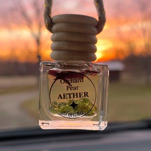 May include: A clear glass car air freshener with a wooden top and a rope for hanging. The label reads "Orchard Pear AETHER". The background shows a colorful sunset.