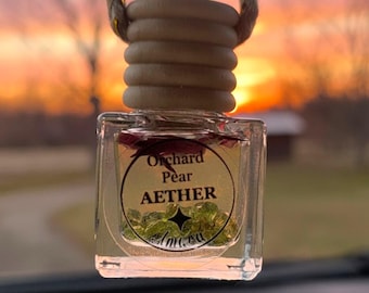May include: A clear glass car air freshener with a wooden top and a rope for hanging. The label reads "Orchard Pear AETHER". The background shows a colorful sunset.