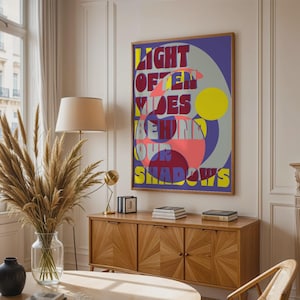 Peut inclure: Une affiche encadrée avec le texte "LIGHT OFTEN HIDES BEHIND OUR SHADOWS" dans un design géométrique coloré. L'œuvre présente un fond violet avec des formes jaunes, roses et grises. L'affiche est exposée dans une pièce avec un meuble en bois et un vase de plantes séchées.