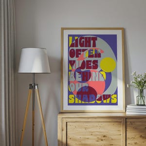 Könnte beinhalten: Ein gerahmter Kunstdruck mit dem Text "LIGHT OFTEN HIDES BEHIND OUR SHADOWS" in farbenfrohen, stilisierten Buchstaben. Das Kunstwerk zeigt ein geometrisches Design mit einem gelben Kreis und rosa und blauen Formen. Der Druck wird auf einem Holzschrank ausgestellt.