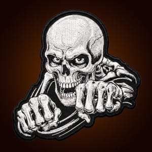 Könnte beinhalten: Ein gestickter Aufnäher mit einem detaillierten weißen Totenkopf mit aggressivem Ausdruck, dargestellt ab der Brust mit skelettierten Händen, die ein Lenkrad umgreifen. Das Design verwendet kontrastreiches weißes Garn auf schwarzem Grund.