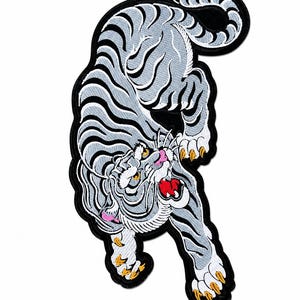 May include: An embroidered patch featuring a stylised tiger with light grey fur and dark grey stripes. The tiger is depicted in a pouncing pose with its mouth open, showing a red tongue and white teeth. It has yellow claws and pink accents.