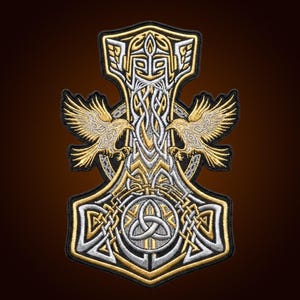 Könnte beinhalten: Ein bestickter Aufnäher mit einem silber- und goldfarbenen Mjolnir-Hammer-Design. Der Hammer ist mit komplizierten Knotenmustern, einem zentralen Triquetra-Symbol und einer stilisierten behelmten Figur verziert, flankiert von zwei goldenen Raben.
