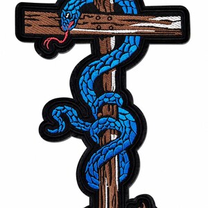 May include: An embroidered patch featuring a bright blue snake with a red tongue coiled around a brown wooden cross. The cross has a textured appearance with white highlights, and the entire design is outlined with a thick black border.