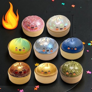 Könnte beinhalten: Acht farbenfrohe, runde Behälter, die Knödeln ähneln, jeweils mit einem lächelnden Gesicht und einem glitzernden oder perlmuttartigen Finish. Die Behälter haben einen Durchmesser von ca. 7 cm und eine Höhe von 7 cm. Die Farben sind Rosa, Weiß, Blau, Grün, Gold und Braun.