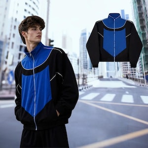 Könnte beinhalten: Schwarz-blaue Trainingsjacke mit weißem Besatz. Die Jacke hat einen Stehkragen und einen durchgehenden Reißverschluss. Die Vorderseite hat eine große blaue Fläche und schwarze Seitenteile. Die Jacke wird von einer Person in einer urbanen Umgebung getragen.