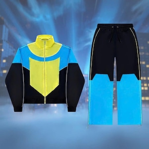 Könnte beinhalten: Ein zweiteiliger Trainingsanzug mit Colorblock-Design. Die Jacke hat eine gelbe Mittelbahn, blaue Oberbahnen und schwarze Ärmel. Die Hose ist schwarz mit blauen Unterbahnen. Beide Teile haben weiße und gelbe Akzente.