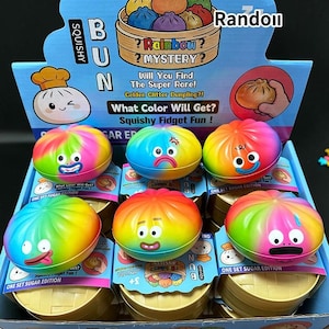 Könnte beinhalten: Eine Präsentation von bunten, matschigen Zappelspielzeugen in Form von Brötchen. Jedes Spielzeug ist ein Regenbogen aus Farben mit einem anderen Gesichtsausdruck. Die Verpackung enthält den Text "Rainbow Mystery?" und "Squishy Fidget Fun!"