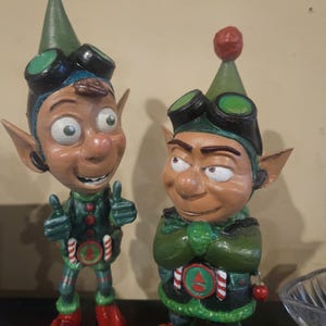 May include: Two whimsical elf figurines. Each elf wears a green and red outfit with a matching hat, goggles, and red shoes. One elf gives a thumbs-up, while the other has arms crossed. The figurines have detailed facial features.