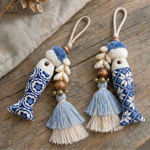 Op de afbeelding: Twee handgemaakte visvormige ornamenten met blauw-witte stoffen lichamen. Elk ornament heeft een kwast, houten kralen, kleine belletjes en een lus om op te hangen. De vissen hebben pompons en schelpaccenten.