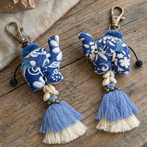 May include: Two blue and white elephant keychains with gold-tone hardware. Each keychain features a fabric elephant, a tassel, and shell accents. The elephants have a floral pattern and are attached to a clasp.