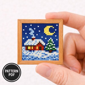 Pode incluir: Uma pequena obra de arte de ponto cruz quadrado numa moldura de madeira. A cena retrata uma cabana coberta de neve, um pinheiro verde e uma lua crescente amarela contra um céu azul escuro. O texto "PATTERN PDF" está num círculo preto.