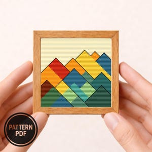 May include: A small, square cross-stitch artwork in a wooden frame. The design features a geometric mountain range in shades of red, orange, yellow, blue, and green against a light yellow background. The words "PATTERN PDF" are in a circular badge.