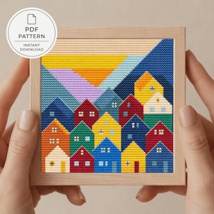 以下が含まれることがあります： 様々な色の家々が並ぶカラフルな村の風景を描いた、額入りのクロスステッチパターン。デザインには夕焼けと山が含まれています。フレームは明るい色の木材で作られています。画像には「PDF PATTERN INSTANT DOWNLOAD」と書かれています。