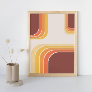 May include: A framed abstract art print with a retro design. The artwork features curved lines in shades of orange, yellow, and brown against a cream background. The frame is made of light-colored wood. A beige vase with dried flowers is in the foreground.
