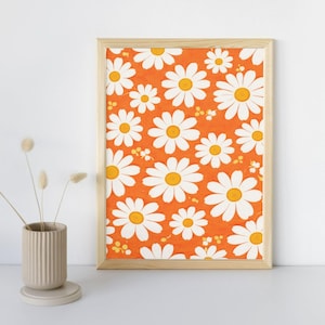 May include: A framed print featuring a repeating pattern of white daisies with yellow centers on an orange background. The artwork is displayed in a light-colored wooden frame, creating a cheerful and vibrant aesthetic.