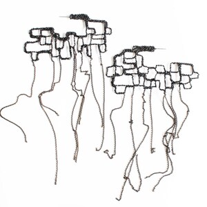 May include: Two abstract, dark metal earrings. Each earring features a geometric, interconnected design with dangling chains. The earrings are set against a white background, highlighting their intricate structure and unique style. The design is modern and artistic.