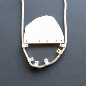 Puede incluir: Un collar con un colgante blanco de forma geométrica y una cadena de cuerda. El colgante está unido a la cadena con pequeños anillos metálicos.