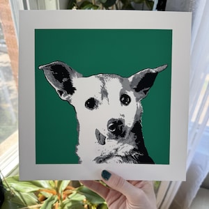 Puede incluir: Una impresión artística cuadrada que presenta una ilustración gráfica estilizada y de alto contraste de un perro con orejas grandes y la lengua ligeramente fuera. El perro está representado en tonos blancos, grises y negros sobre un fondo verde sólido.