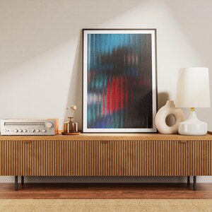 May include: A wooden media console with a slatted front, holding a framed abstract art print with blue, red, and black colors. A silver stereo receiver, a white lamp, and a ceramic vase are also on the console.