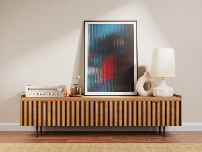 May include: A wooden media console with a slatted front, holding a framed abstract art print with blue, red, and black colors. A silver stereo receiver, a white lamp, and a ceramic vase are also on the console.