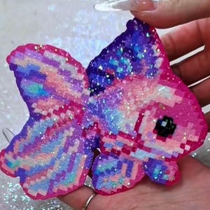 May include: A pixelated, glitter-covered goldfish in shades of pink, purple, and blue. The fish has a black eye and is held in a hand. The design is reminiscent of a galaxy.