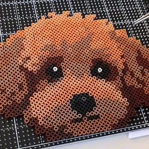 May include: A pixel art dog's face created from small, cylindrical beads in shades of brown. The dog has a black nose and white eyes. The artwork is set against a black and white grid, with tweezers in the background.