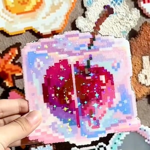 May include: A pixelated coaster in a square shape, depicting a red apple with a straw. The coaster has a pink, blue, and white colour scheme with glitter accents. Other pixel art coasters are visible in the background.