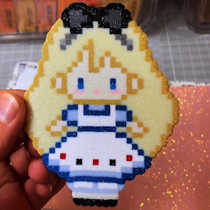 May include: A pixelated Alice in Wonderland design, featuring a blonde-haired girl in a blue and white dress with a black bow. The artwork is made of small, colourful beads, with a glittery finish. The edges are outlined in a golden colour.