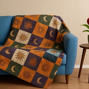 May include: A colorful throw blanket with a celestial design featuring suns and moons in a checkerboard pattern. The blanket is draped over a blue sofa. A red flower in a green pot sits on a wooden side table.