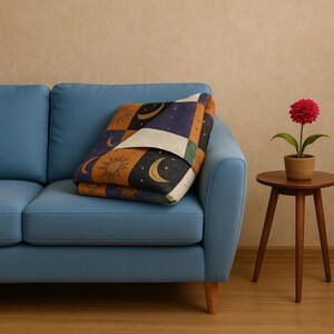 May include: A folded blanket with a celestial pattern of suns, moons, and stars in orange, navy, and black. The blanket rests on a light blue sofa. A small, round wooden side table holds a potted red flower.
