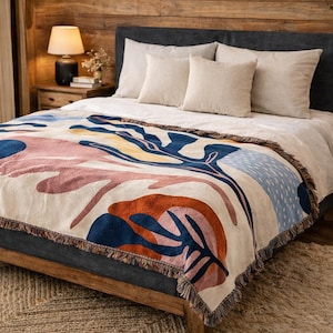 Puede incluir: Una manta de color crema con un diseño abstracto en tonos azules, naranjas, rosas y amarillos. La manta tiene un borde con flecos y se muestra en una cama con almohadas de color neutro.