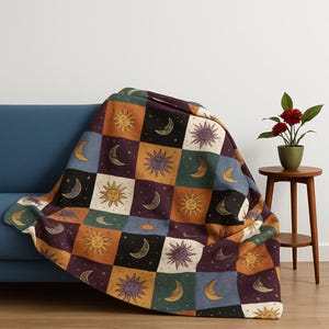 May include: A patterned throw blanket with a patchwork design featuring suns, moons, and stars in shades of orange, purple, and blue. The blanket is draped over a blue sofa, creating a cosy and decorative look.