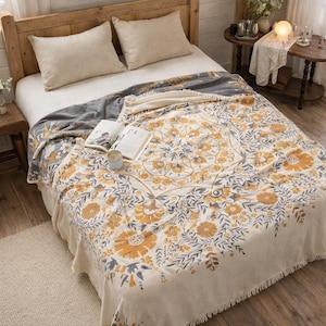 May include: A bedspread with a floral pattern in beige, grey, and yellow. The bed is made with two beige pillows. The blanket has a large, circular floral design. A book and candle are on the bed.