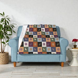 May include: A patterned throw blanket with a celestial theme, displaying squares with suns, moons, and stars in shades of purple, orange, and blue. The blanket is draped over a light blue armchair, adding a decorative and cosy element.