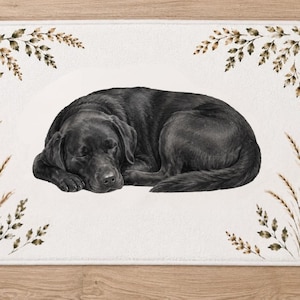 Puede incluir: Una alfombrilla de baño rectangular con esquinas redondeadas que presenta una ilustración central de un perro Labrador negro durmiendo. El fondo es de color blanco roto, decorado con ramitas de hojas y hierbas marrones y beige en las cuatro esquinas.