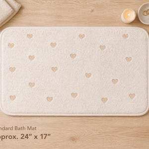 Op de afbeelding: Crèmekleurige rechthoekige badmat met geborduurd hartpatroon. De mat meet ongeveer 61 cm x 43 cm. De mat ligt op een houten oppervlak, met een kaars, een kleine kom en opgevouwen handdoeken.