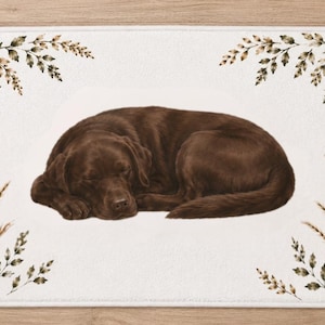Op de afbeelding: Een rechthoekige badmat met een witte achtergrond en een centrale illustratie van een slapende chocoladebruine Labrador retriever. De hoeken zijn versierd met botanische illustraties van bruine en groene bladeren en hoge grassprieten.