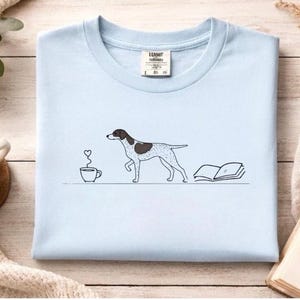 May include: Light blue crew neck t-shirt featuring a line art illustration of a dog with a brown and white pattern, a coffee cup with a heart steam, and an open book. The shirt is a soft, comfortable material.