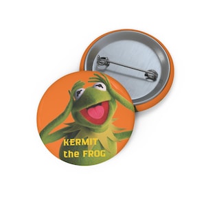 May include: Two orange and silver pinback buttons. One button features a green Kermit the Frog character with his hands on his head and his tongue out. The text "KERMIT the FROG" is printed on the button.
