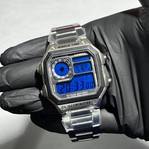 Peut inclure: Une montre numérique argentée avec un bracelet à maillons en acier inoxydable. Le cadran carré présente un affichage bleu avec le texte WORLD TIME, CASIO, WR100M, 10 YEAR BATTERY et ILLUMINATOR. La montre inclut un sous-cadran analogique.