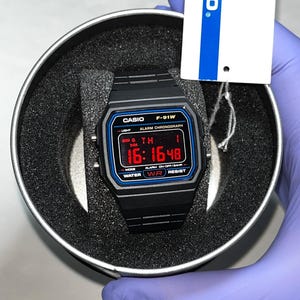 Op de afbeelding: Een zwart Casio F-91W digitaal horloge met een zwarte kunsthars band en een rechthoekige wijzerplaat met een rood digitaal display dat 16:16:48 aangeeft. Het horloge heeft blauwe accenten rond het scherm en zit in een ronde houder met een wit label.
