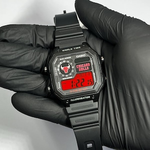 Op de afbeelding: Een zwart Casio digitaal horloge met een harsband, voorzien van een vierkante wijzerplaat met een rood display. Het scherm toont het Chicago Bulls-logo, de tekst Chicago Bulls en de tijd 1:22:15. Het horloge bevat World Time, 5 Alarms en Illuminator.