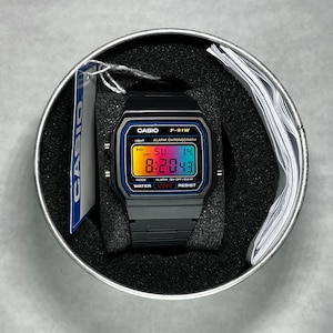 Puede incluir: Un reloj digital Casio F-91W negro con una pantalla de colores arcoíris, que presenta el texto CASIO, F-91W, ALARM CHRONOGRAPH y WATER RESIST. El reloj está alojado en una lata redonda de metal con espuma negra y una etiqueta azul colgante.