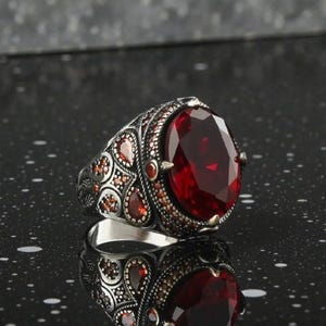 May include: A silver ring with a large, oval-cut, deep red gemstone. The ring has detailed patterns around the central stone and along the band, with smaller red stones adding to the design. The ring is shown on a reflective surface.