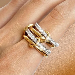May include: A gold-toned ring with a unique design, featuring two parallel bands adorned with sparkling, clear gemstones. The bands are connected by a knot-like structure, creating an elegant and eye-catching piece of jewelry.