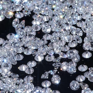 Könnte beinhalten: Eine Ansammlung zahlreicher kleiner, klarer, facettierter runder Edelsteine, die auf einer dunklen Oberfläche verstreut sind. Jeder Stein reflektiert das Licht und erzeugt helle weiße Glanzlichter sowie subtile prismatische Funken.