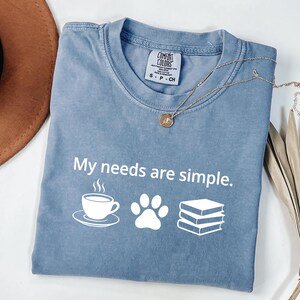 May include: A blue short-sleeved t-shirt with a crew neckline and a white graphic print that reads My needs are simple. The design includes icons of a steaming coffee cup, a paw print, and a stack of three books. The shirt has a Comfort Colors label.
