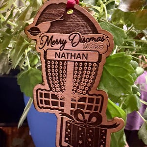 K&ouml;nnte beinhalten: Weihnachtsbaumschmuck aus Holz mit einem Disc-Golf-Korb-Design. Der Schmuck tr&auml;gt den Text "Merry Discmas 2025" und den Namen "Nathan". Ein rotes Band ist zum Aufh&auml;ngen befestigt. Der Schmuck ist braun mit einem Geschenkbox-Design.